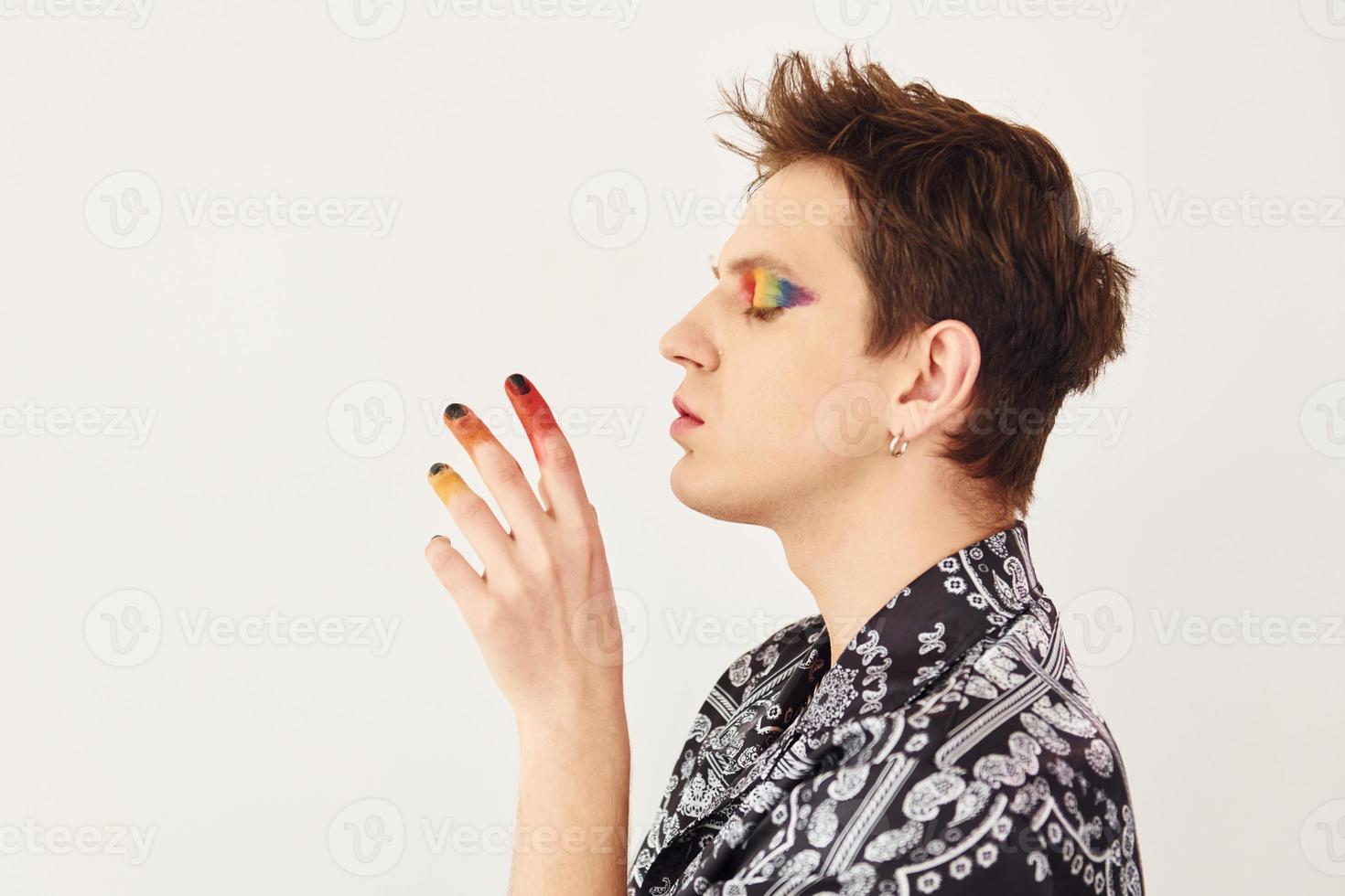 Young gay man is standing in the studio and posing for a camera. Multicolored make up on face fingers photo