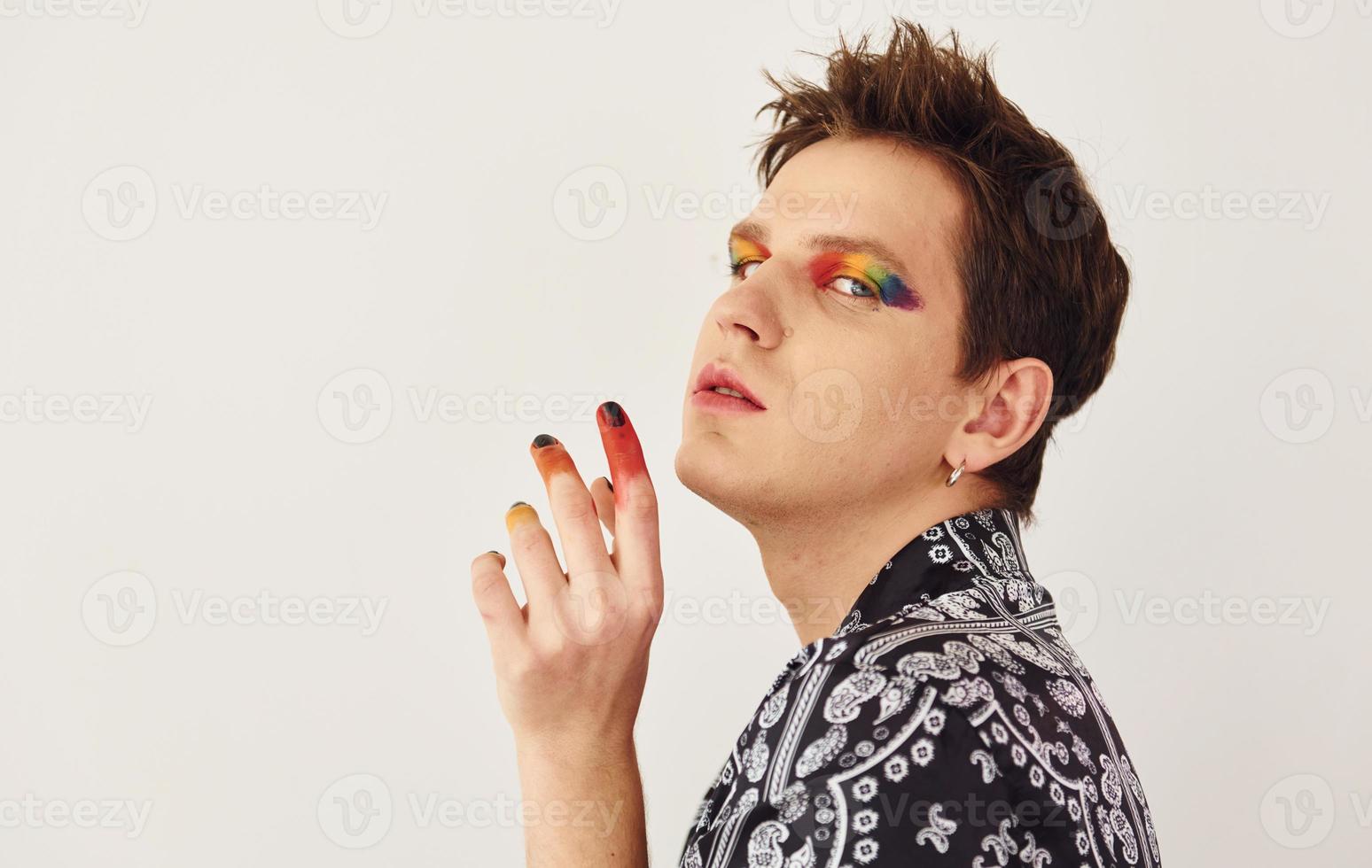 Young gay man is standing in the studio and posing for a camera. Multicolored make up on face fingers photo