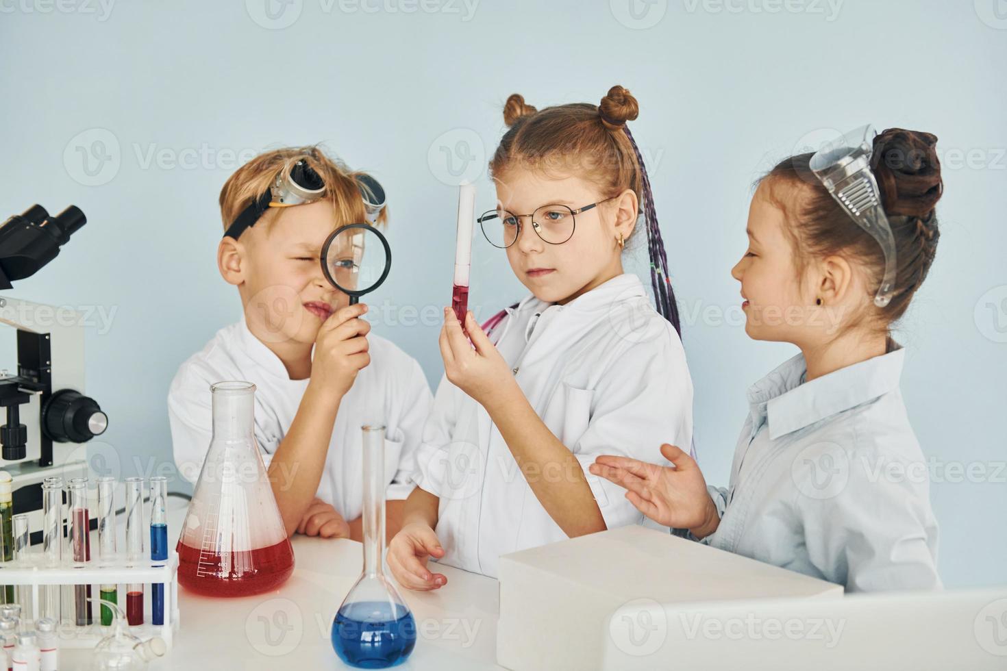 Standing by the table with test tubes. Children in white coats plays a scientists in lab by using equipment photo