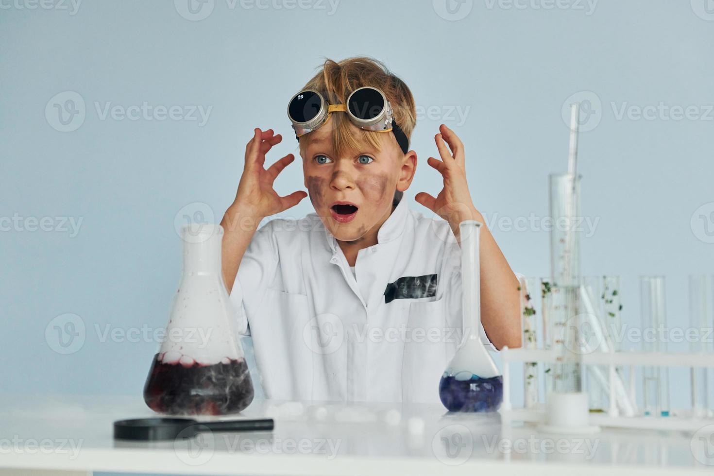 Scared little boy in coat playing a scientist in lab by using equipment photo