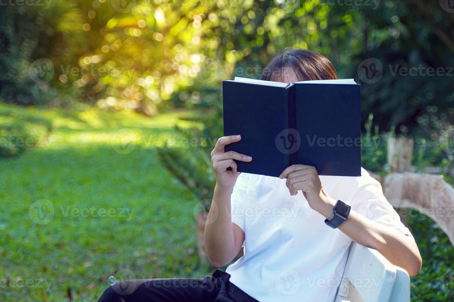 Asian woman holding a book in front of her face while sitting and reading park bench corner. Concept. Asian woman doing outdoor activities, such as reading books, working, having a picnic with family photo