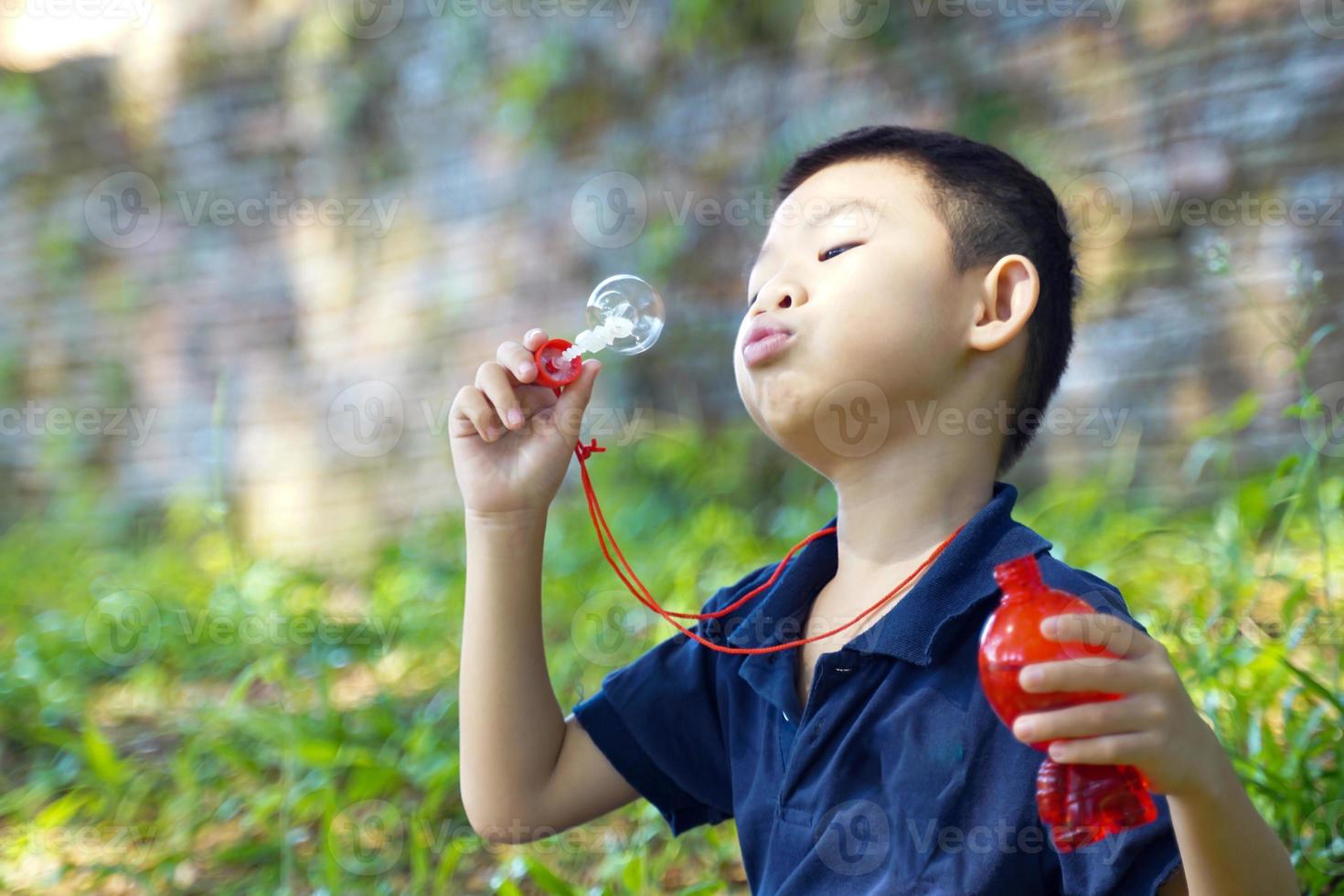Asian boy sitting on the park grass Playing in a toy bubble blower. Concept. Playing in the outdoors, children playing outdoors, children's toys, children's leisure activities. photo