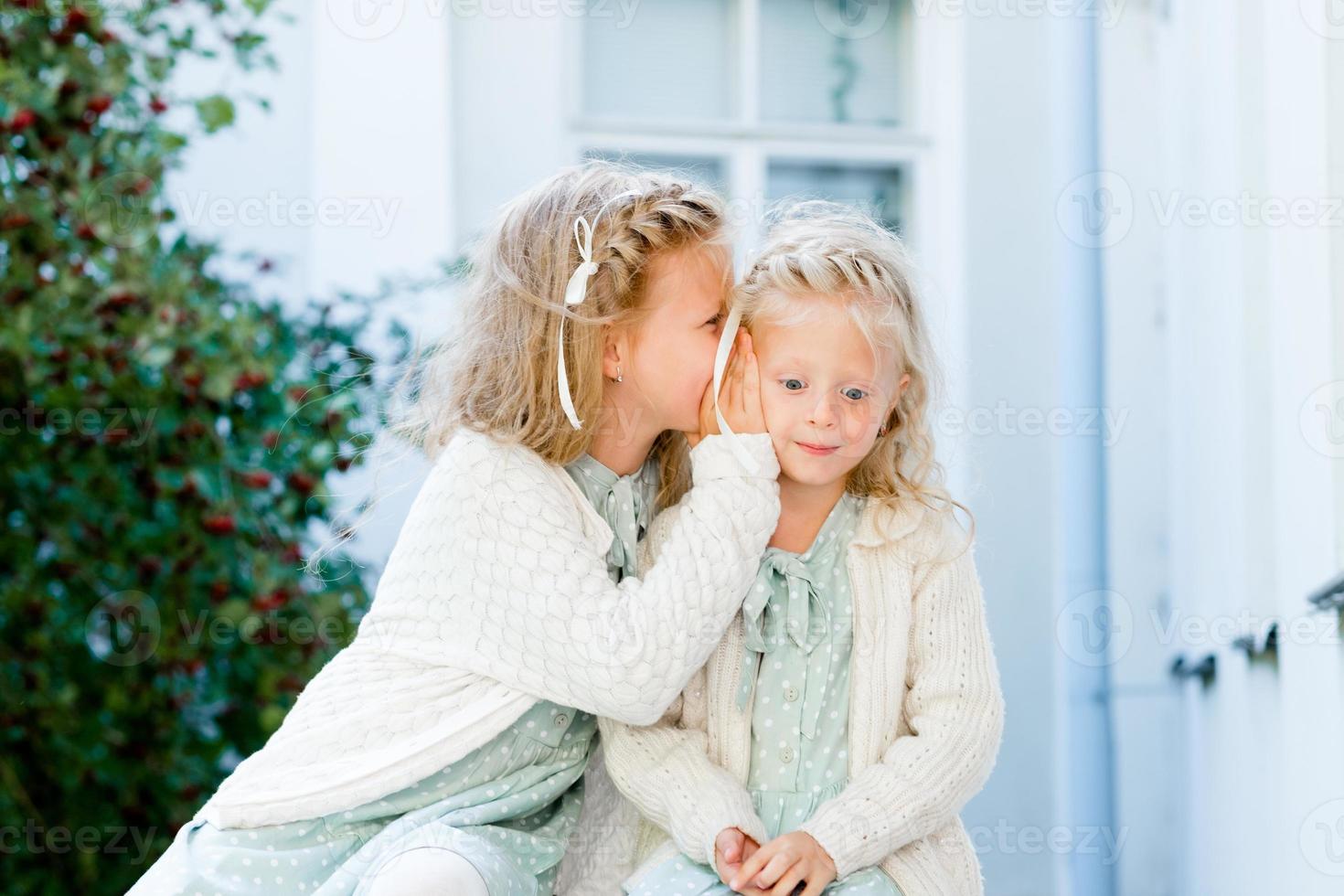 Two little cute girls are sitting in the garden and sharing secrets. One girl whispers something in another's ear. The two sisters have a close relationship. photo