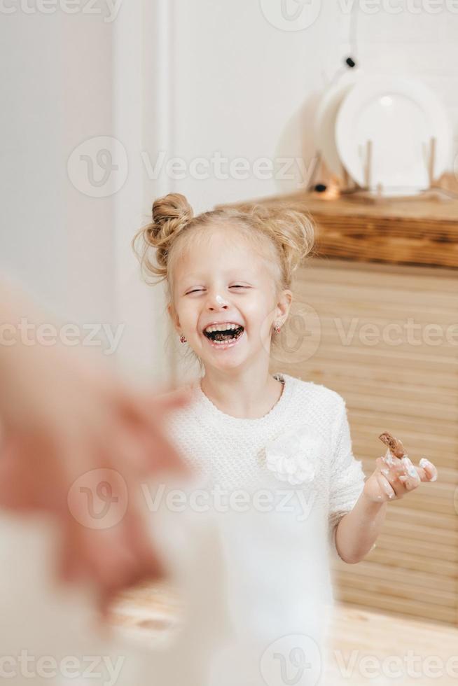 blonde girl paints gingerbread cookies with white cream. Child decorates Christmas cookies with icing on a wooden table in the kitchen. Fooling around with children in the kitchen. photo