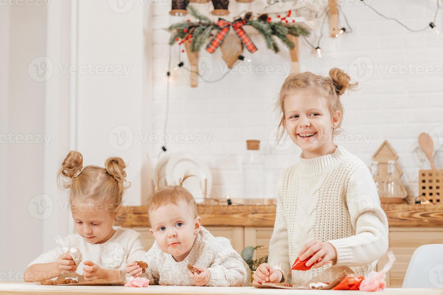 3 blonde girls decorate Christmas cookies with icing on a wooden table in the kitchen. The girlfriends are cooking together in the kitchen with Christmas decor. photo