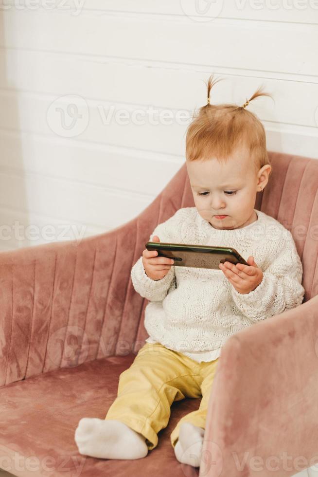 a little girl with ponytails sits on a chair with a phone in her hands. Gadgets from childhood photo