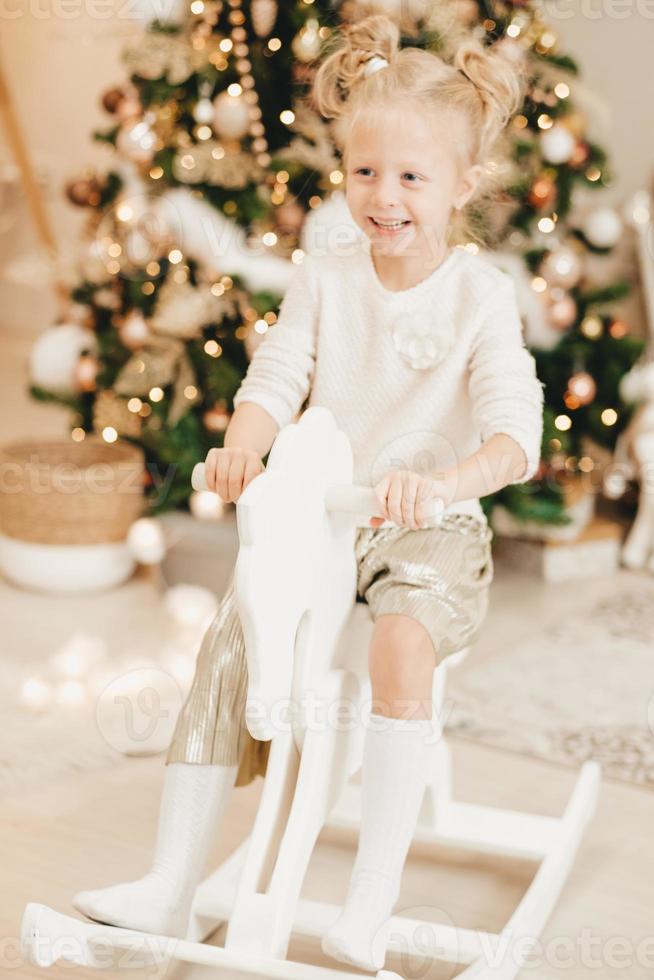 A blonde girl in a sweater rides a wooden horse at home against the background of the Christmas tree. Cute little girl playing at home on the background of garlands. photo