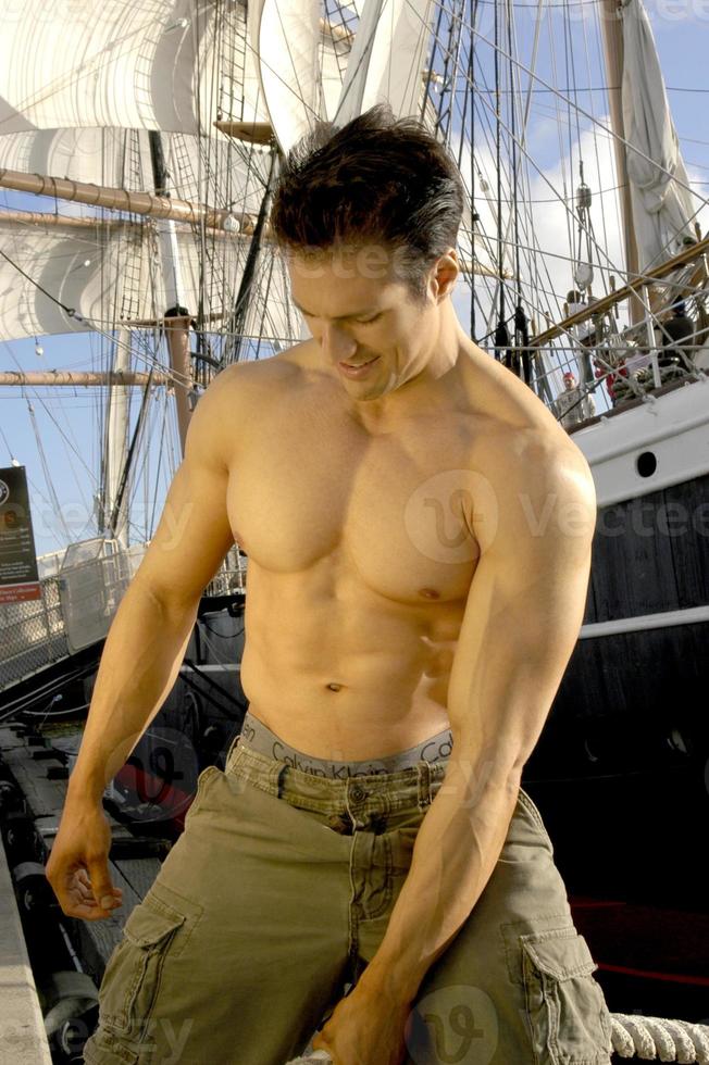 Handsome shirtless muscular captain poses by a sailboat pulling on the ropes with sails drawn. photo