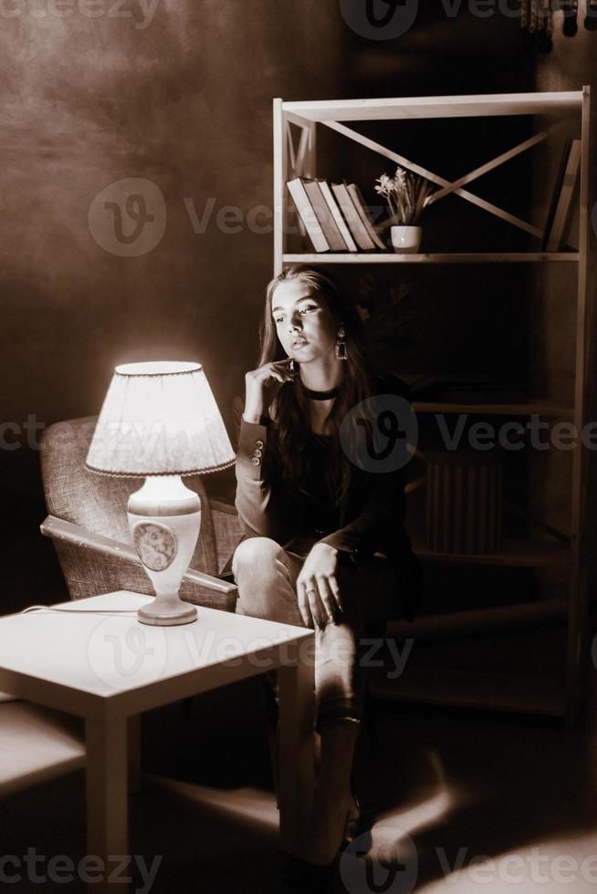 An adult girl sits in a chair near the table. Black and white photo of a girl in a room.