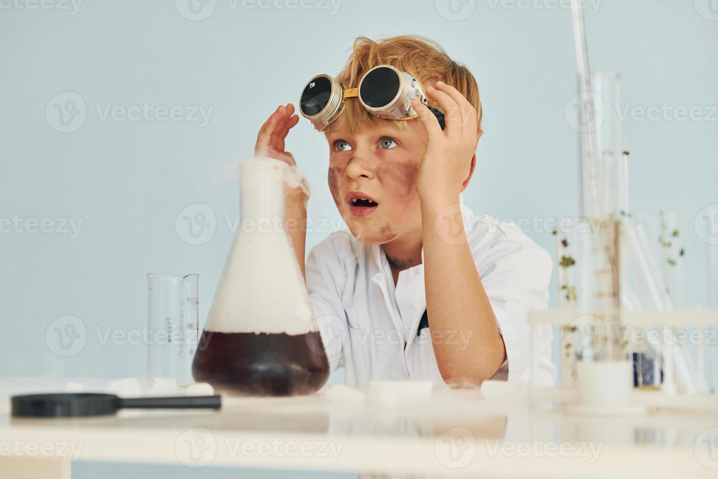 Scared little boy in coat playing a scientist in lab by using equipment photo