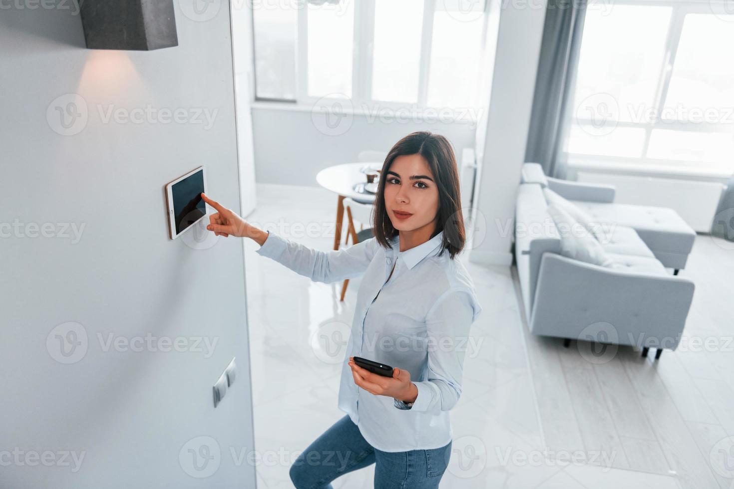 Uses tablet that is mounted in the wall. Young woman is indoors in smart house room at daytime photo