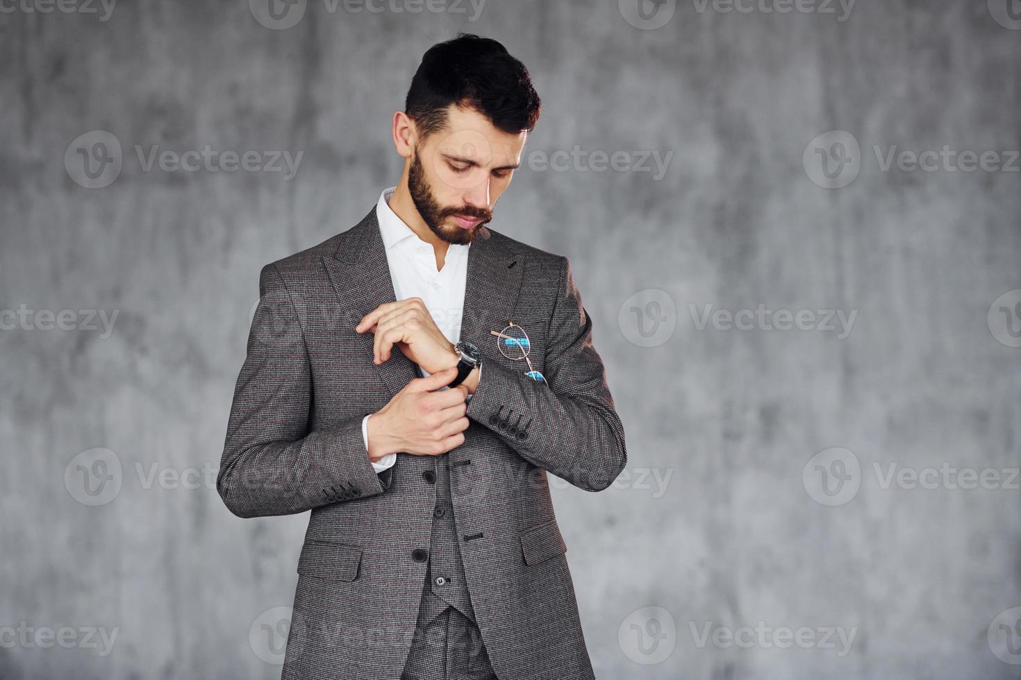 Standing against grey background. Young stylish businessman in suit indoors. Conception of success photo