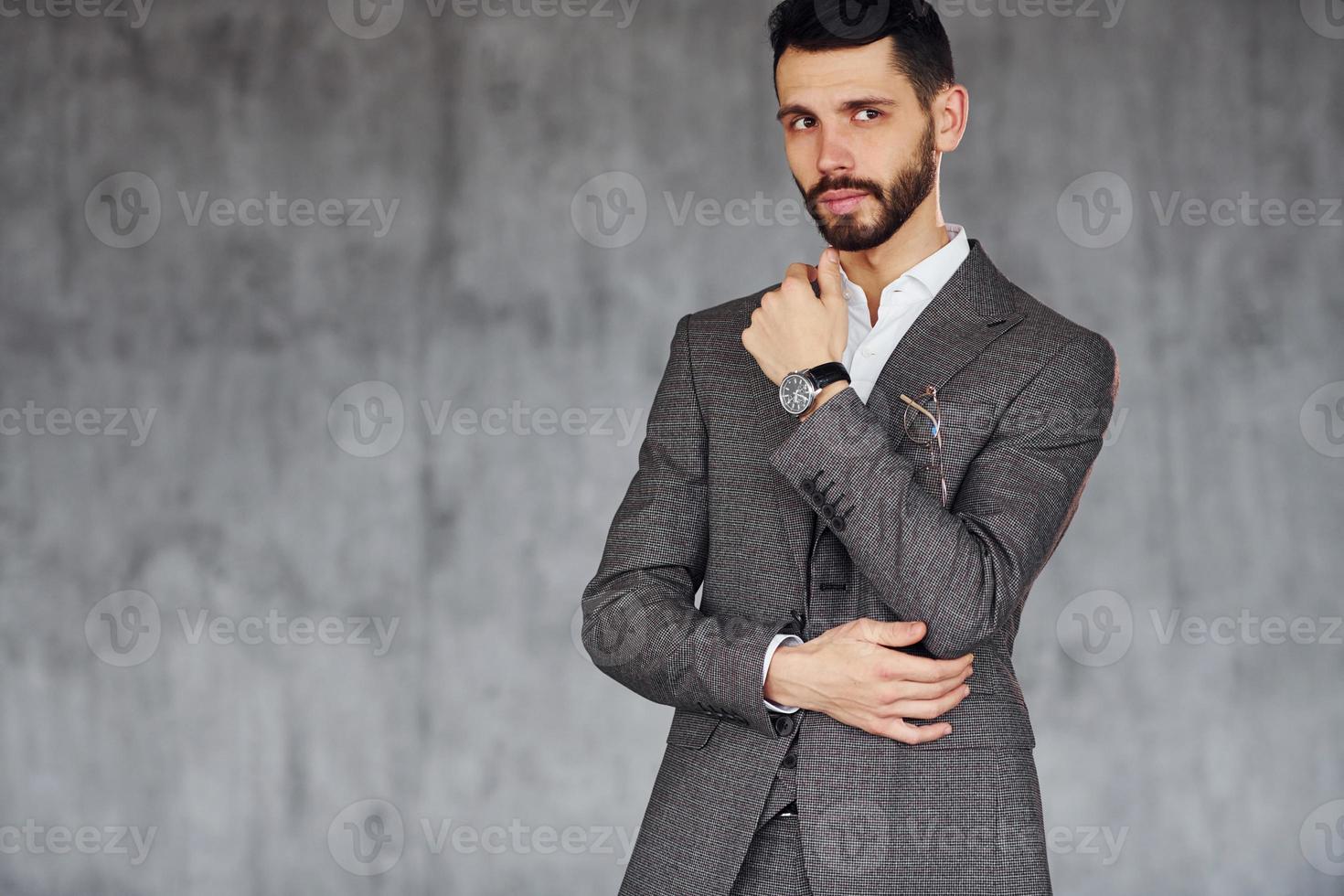 Standing against grey background. Young stylish businessman in suit indoors. Conception of success photo