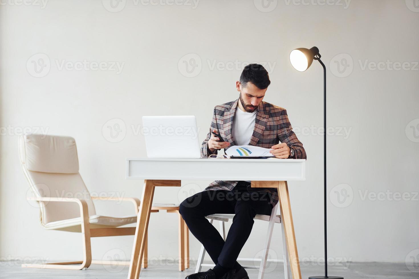 Sits by table in office. Young stylish businessman in suit indoors. Conception of success photo