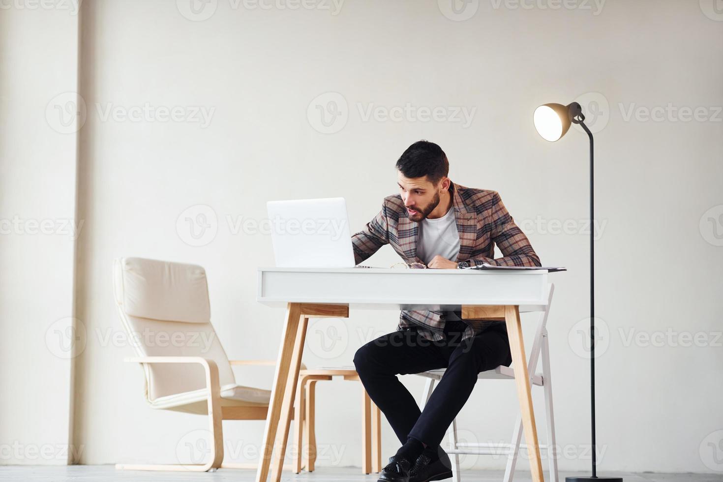 Sits by table in office. Young stylish businessman in suit indoors. Conception of success photo
