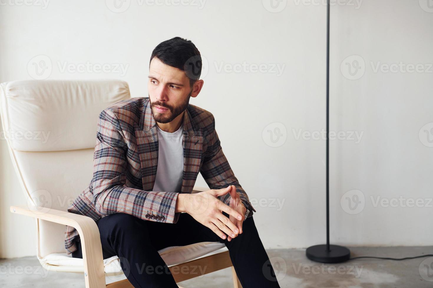 Sits on the chair. Young stylish businessman in suit indoors. Conception of success photo