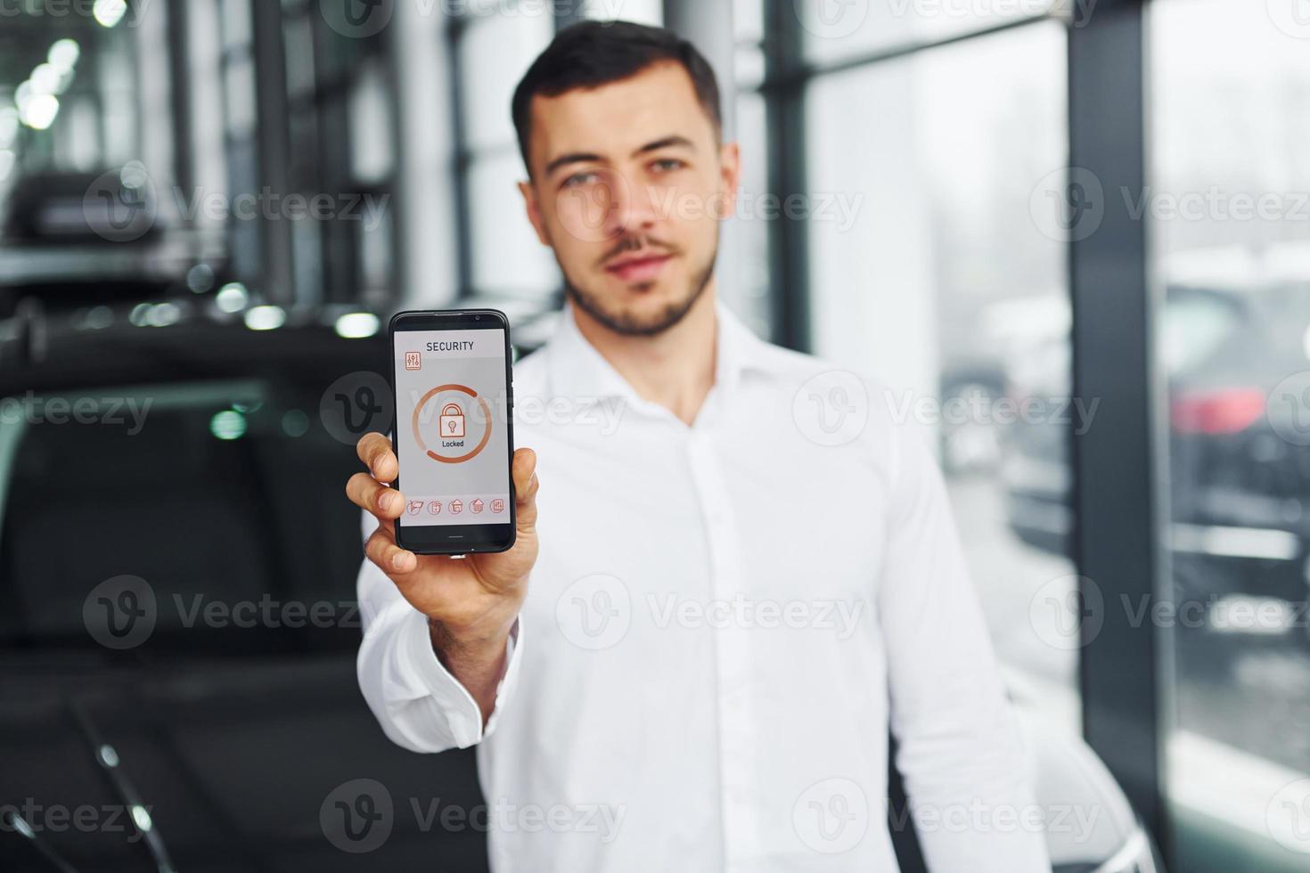 Holds phone with labels and icons. Young man in white shirt is indoors with modern new automobile. Remote control photo