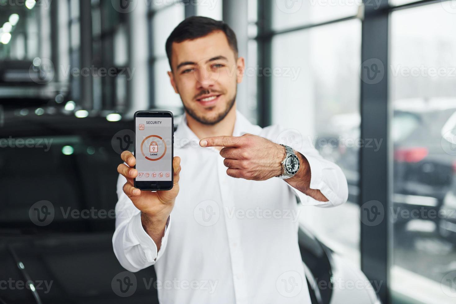 Holds phone with labels and icons. Young man in white shirt is indoors with modern new automobile. Remote control photo