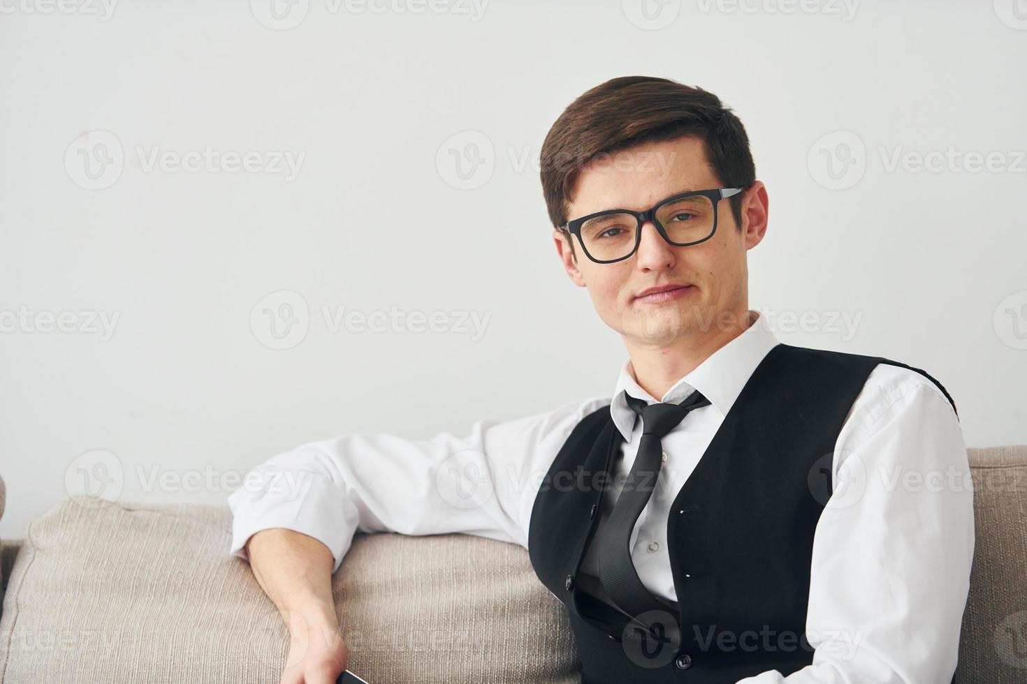 Young man in formal clothes sits on the sofa indoors against white wall photo