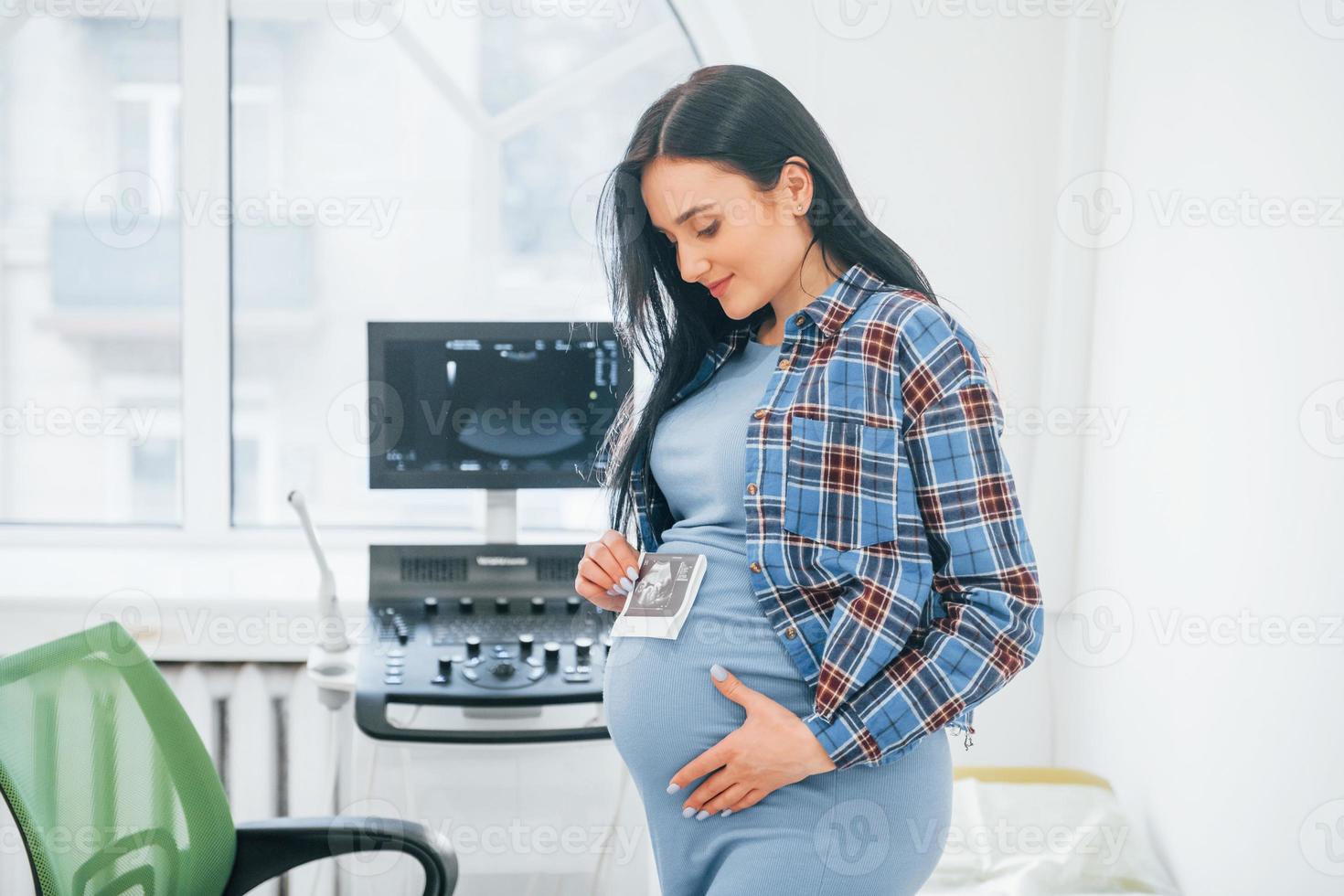 Holds results of ultrasonic scan. Pregnant woman is in the clinic in cabinet and have health check photo