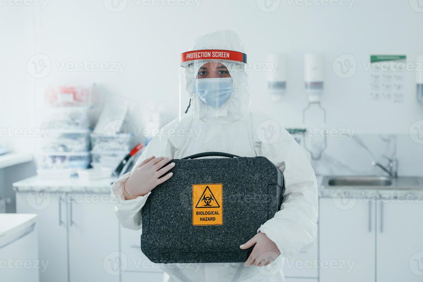 Holds case with biological hazard. Female medical worker in protective uniform works on COVID-19 vaccine photo