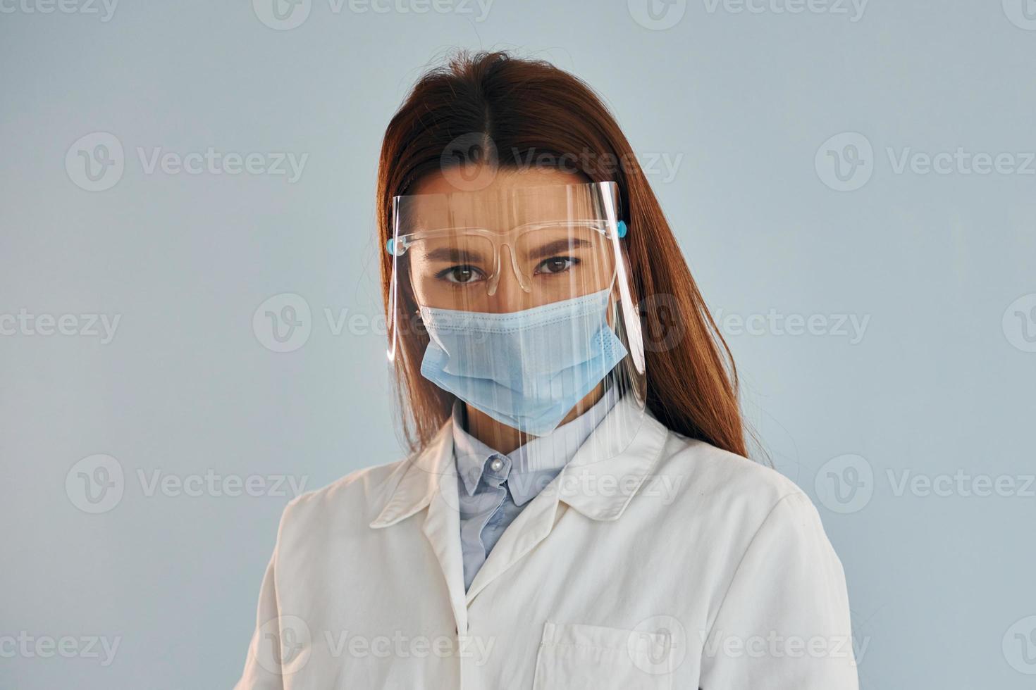 Young female doctor in white coat is standing indoors against blue background photo
