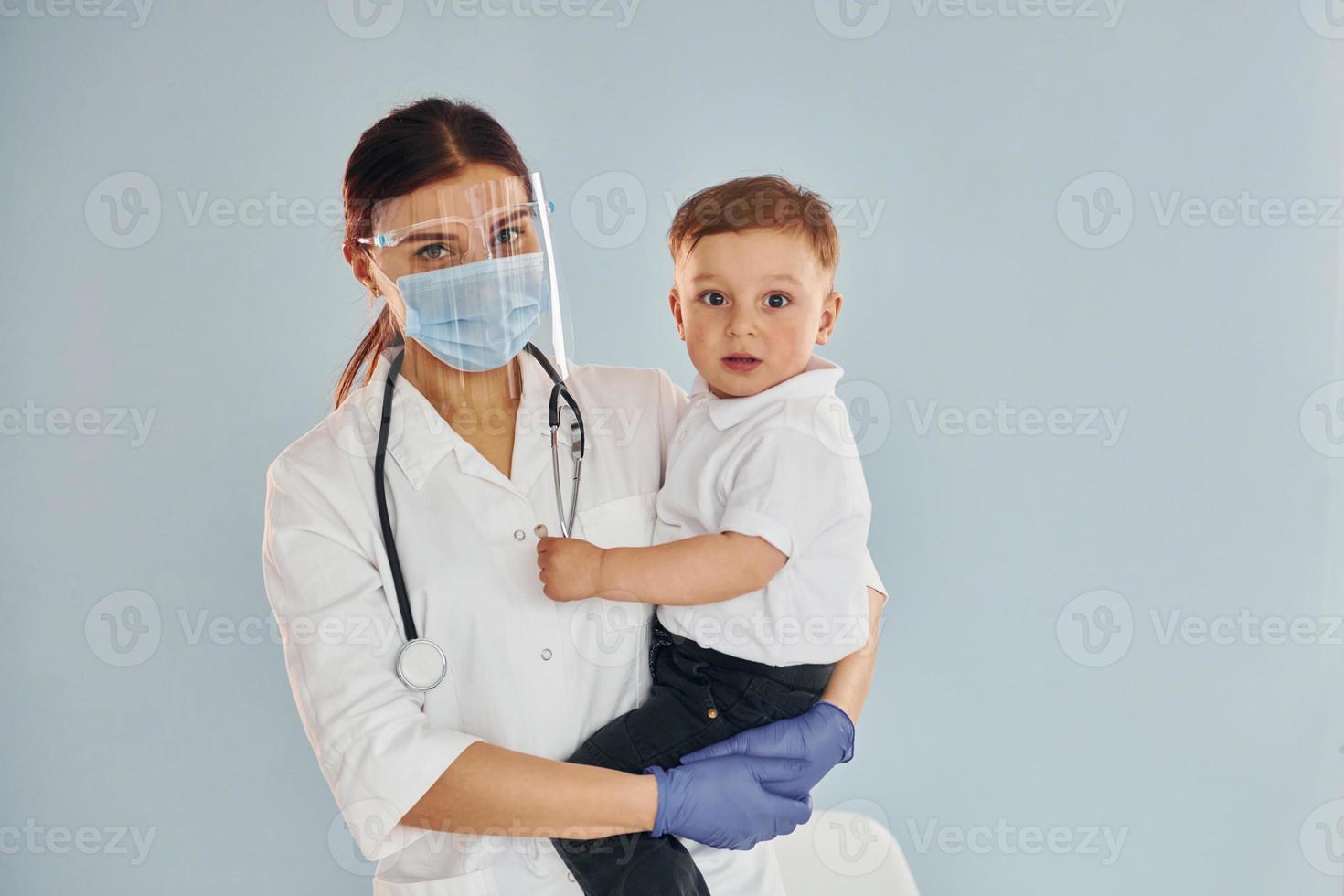 Young nurse in white coat and with stethoscope holds little boy in hands photo