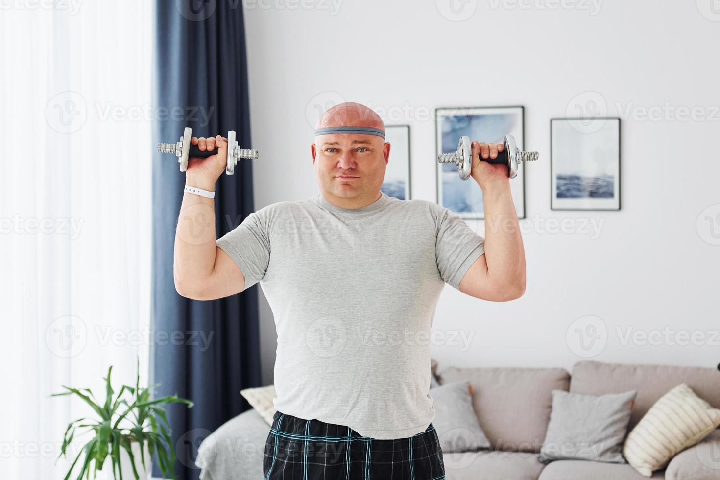 Uses dumbbells. Funny overweight man in casual clothes is indoors at home photo