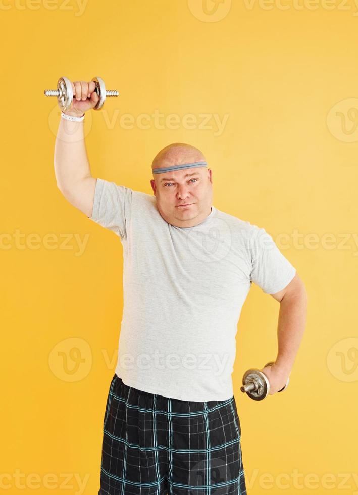 Exercises by using dumbbells. Funny overweight man in sportive head tie is against yellow background photo