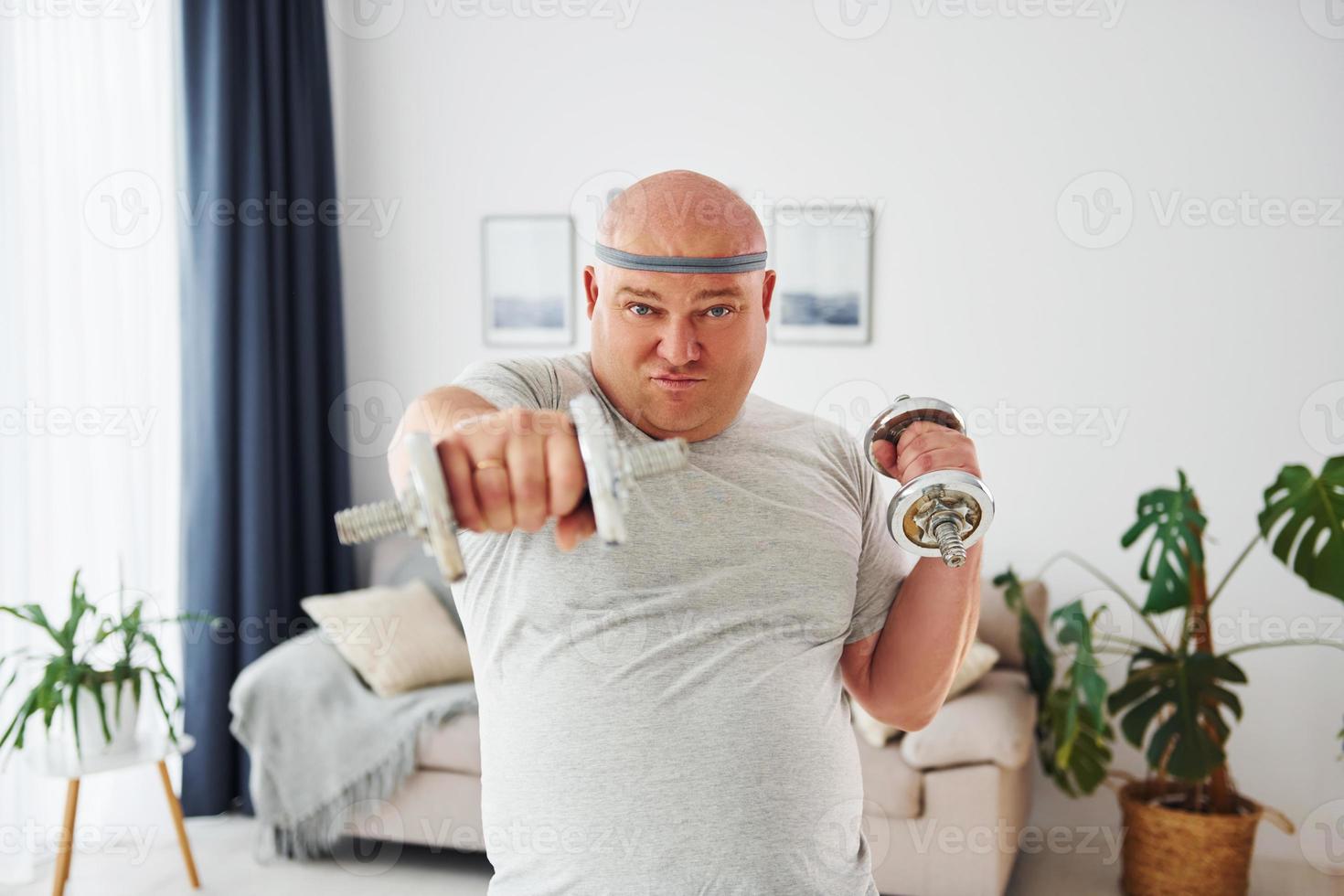 utiliza mancuernas. un hombre gracioso con sobrepeso vestido con ropa informal está en el interior de su casa foto