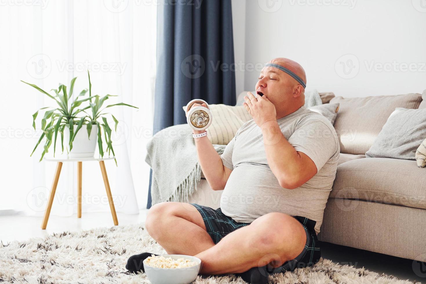 Eats and does exercises. Funny overweight man in casual clothes is indoors at home photo