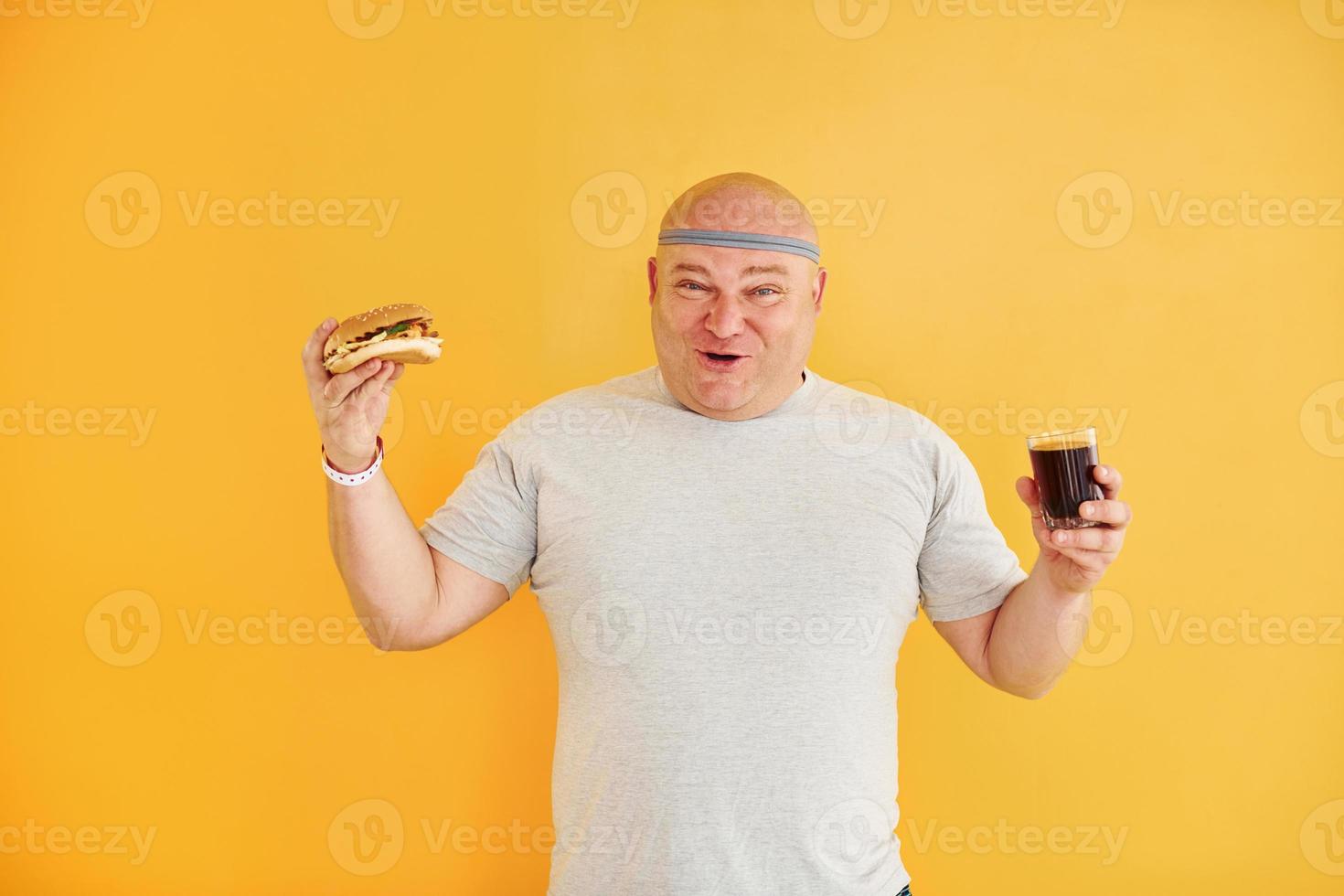 With fast food. Funny overweight man in sportive head tie is against yellow background photo