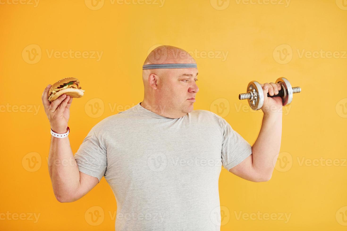 Holds hamburger and dumbbell. Funny overweight man in sportive head tie is against yellow background photo