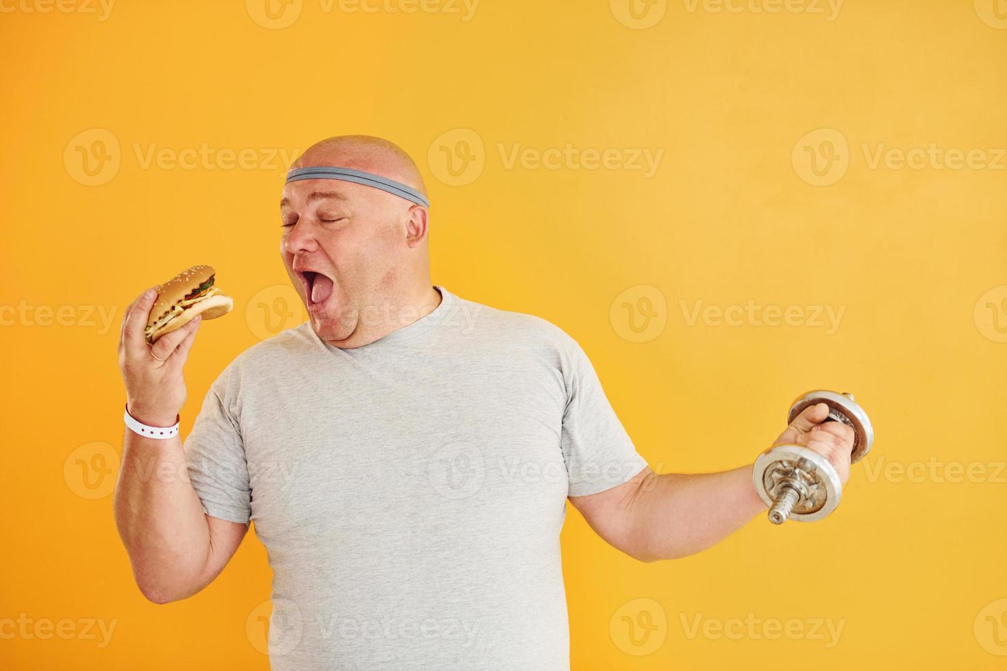 Holds hamburger and dumbbell. Funny overweight man in sportive head tie is against yellow background photo