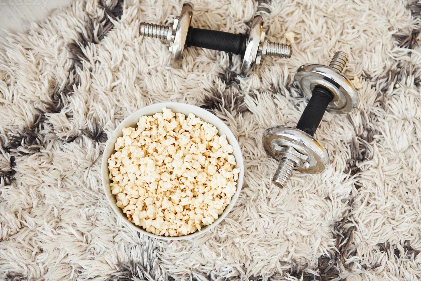 Top view of popcorn in white plate and dumbbells that lying down on the floor photo