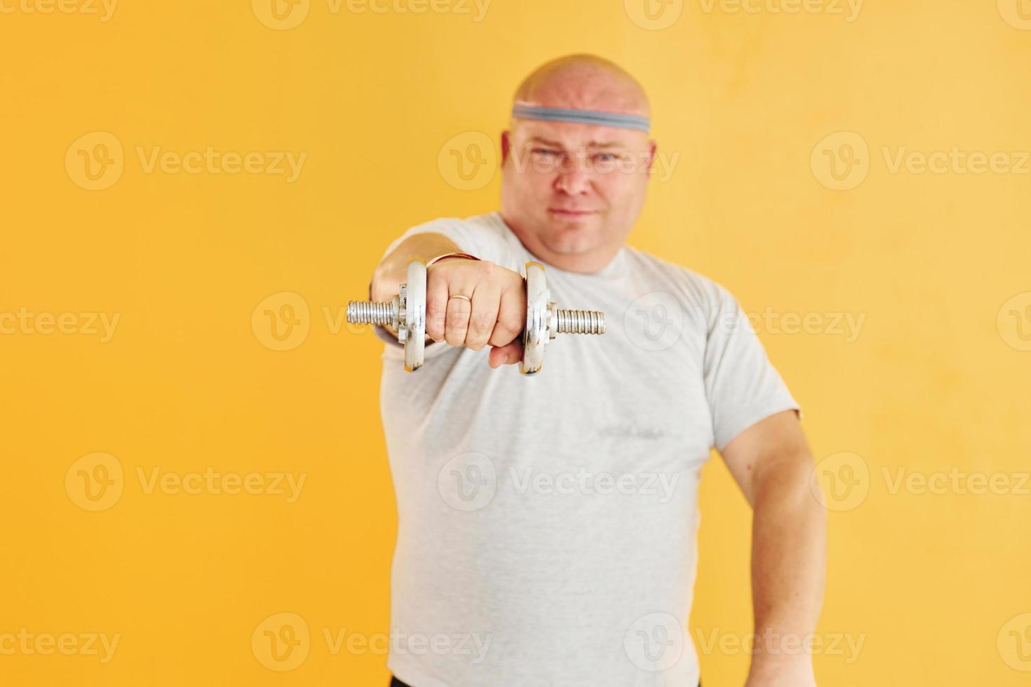 Exercises by using dumbbells. Funny overweight man in sportive head tie is against yellow background photo