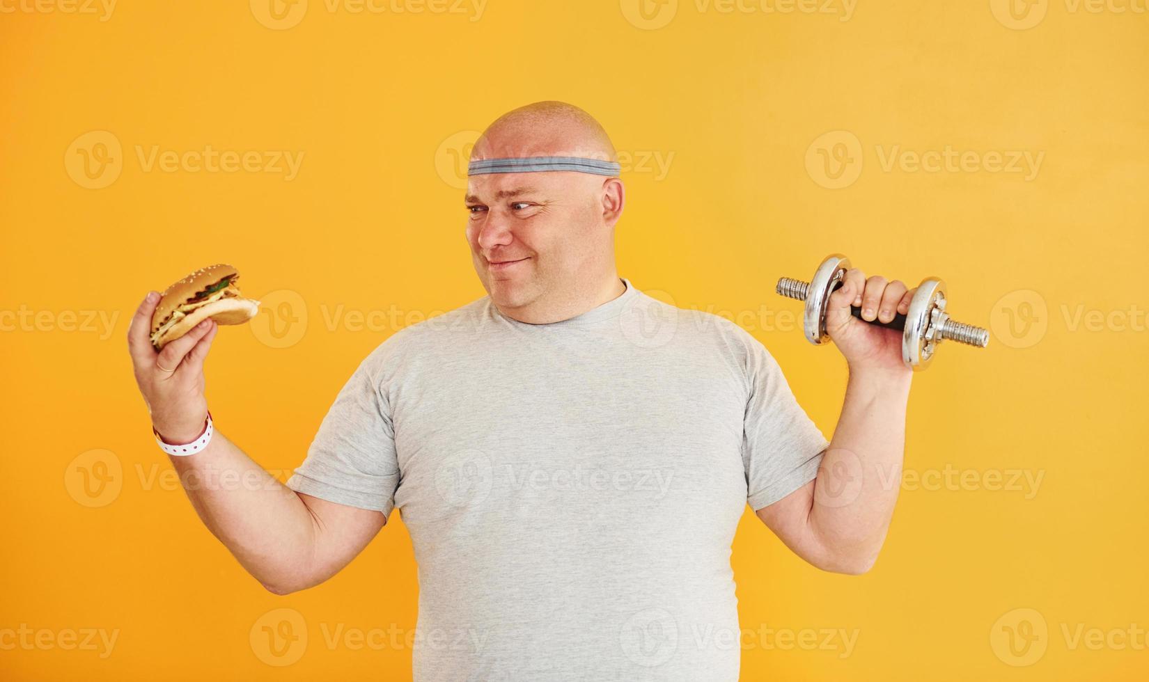 Holds hamburger and dumbbell. Funny overweight man in sportive head tie is against yellow background photo