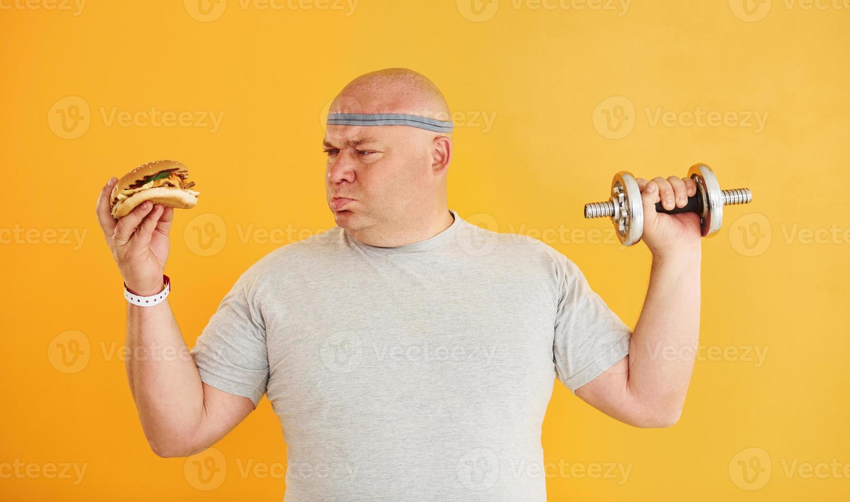 Holds hamburger and dumbbell. Funny overweight man in sportive head tie is against yellow background photo