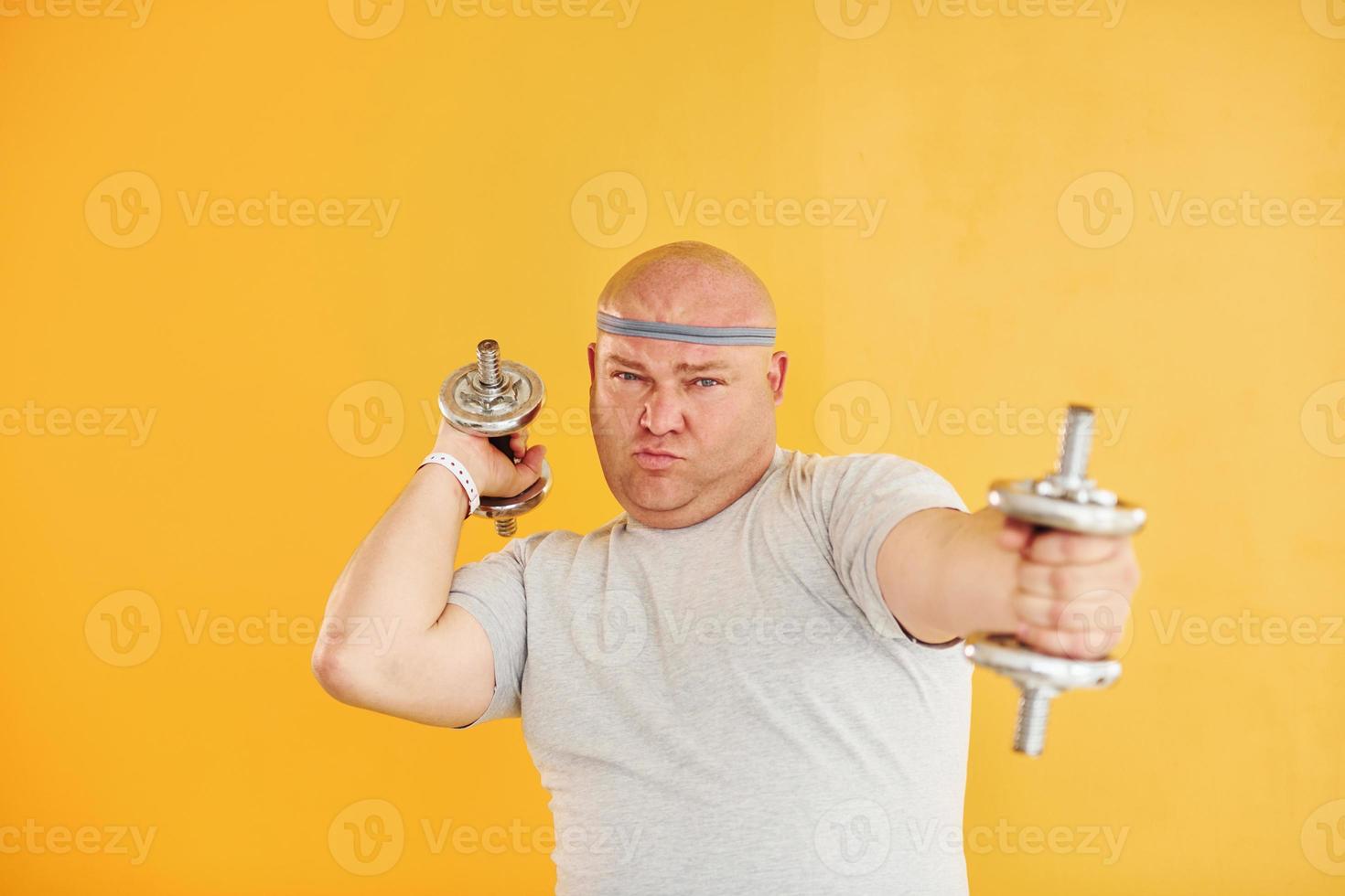 Exercises by using dumbbells. Funny overweight man in sportive head tie is against yellow background photo