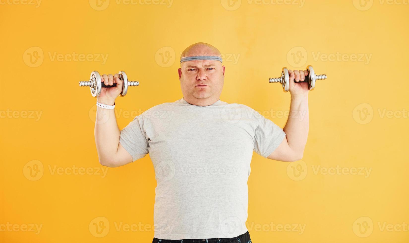Exercises by using dumbbells. Funny overweight man in sportive head tie is against yellow background photo