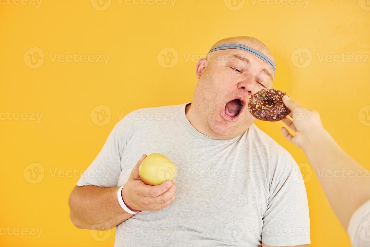 manzana o galleta. el hombre gracioso con sobrepeso en la corbata de cabeza deportiva está contra el fondo amarillo foto