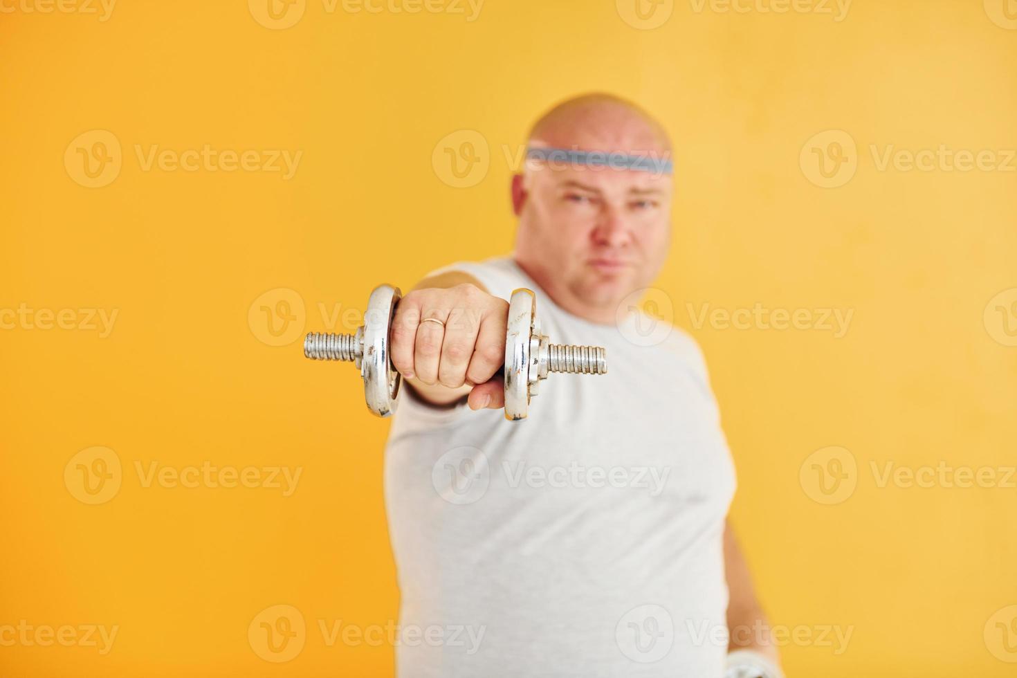 Exercises by using dumbbells. Funny overweight man in sportive head tie is against yellow background photo