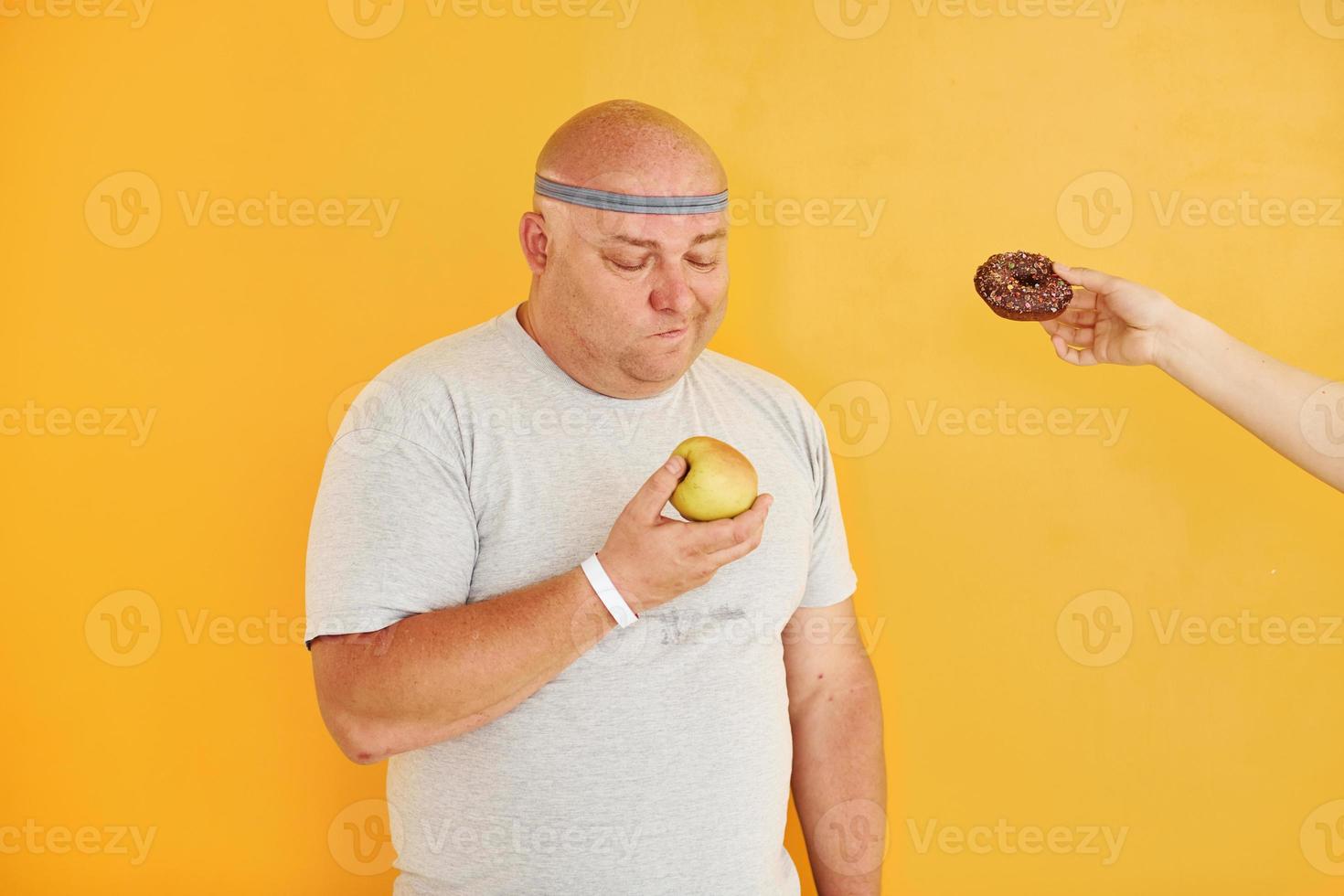 manzana o galleta. el hombre gracioso con sobrepeso en la corbata de cabeza deportiva está contra el fondo amarillo foto