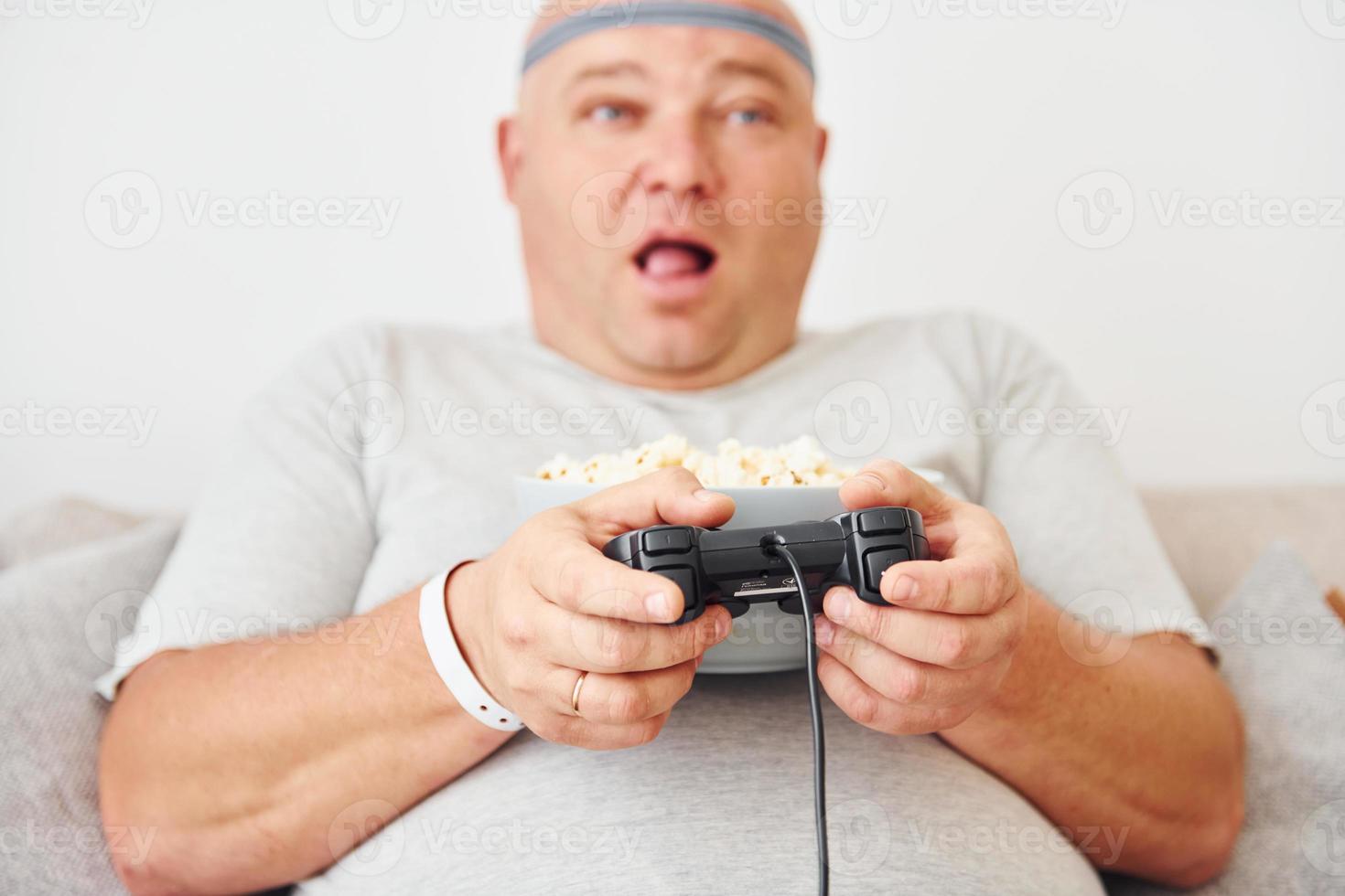 Plays video game. Funny overweight man in casual clothes is indoors at home photo