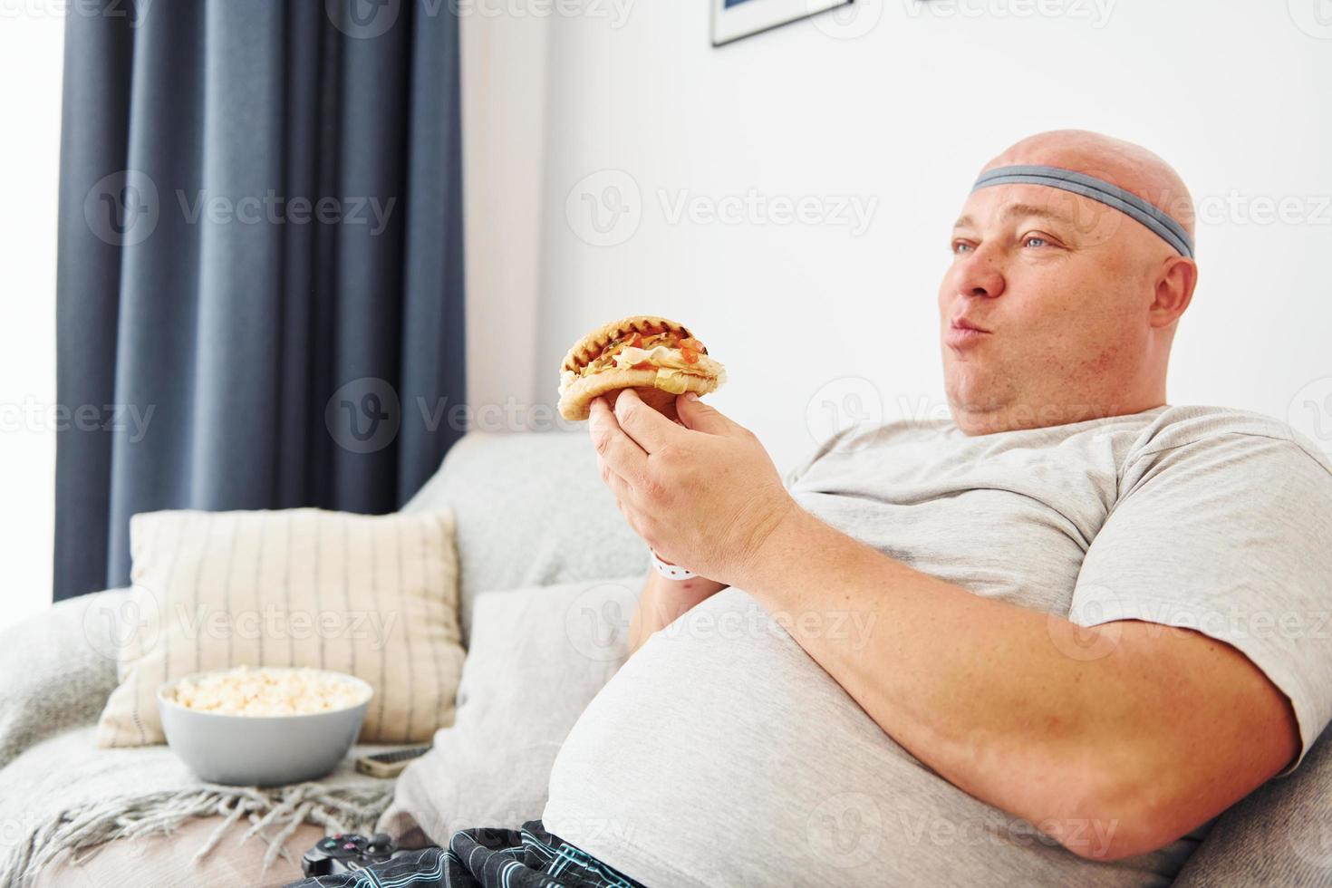 Sits on the sofa with popcorn and hamburger. Funny overweight man in casual clothes is indoors at home photo