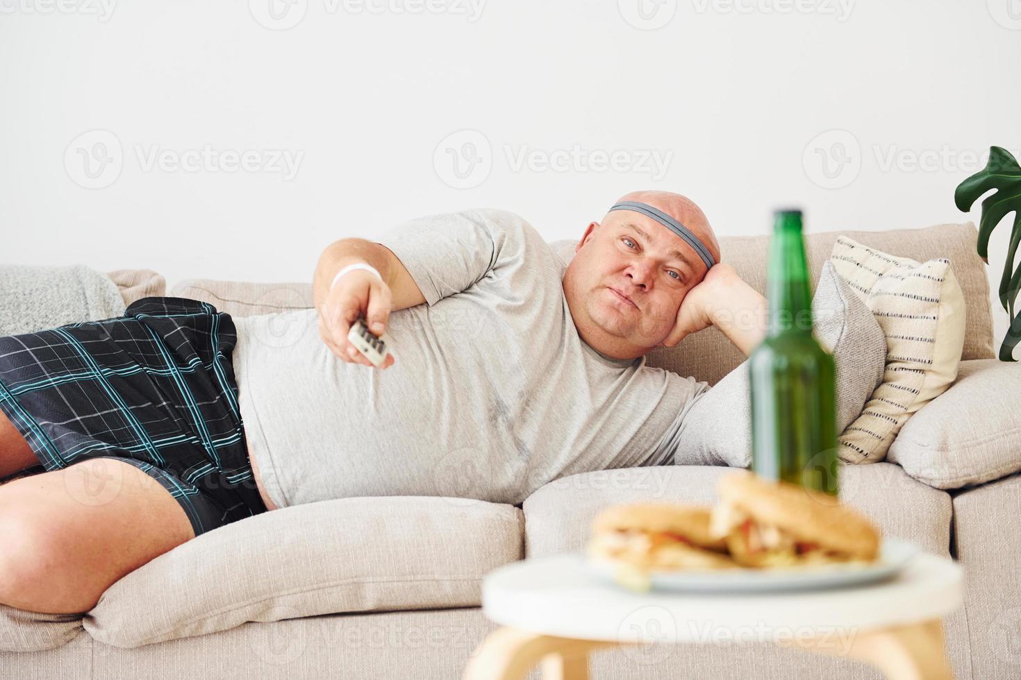 hamburguesas y cerveza. hombre acostado en el sofá. un hombre gracioso con sobrepeso vestido con ropa informal está en el interior de su casa foto