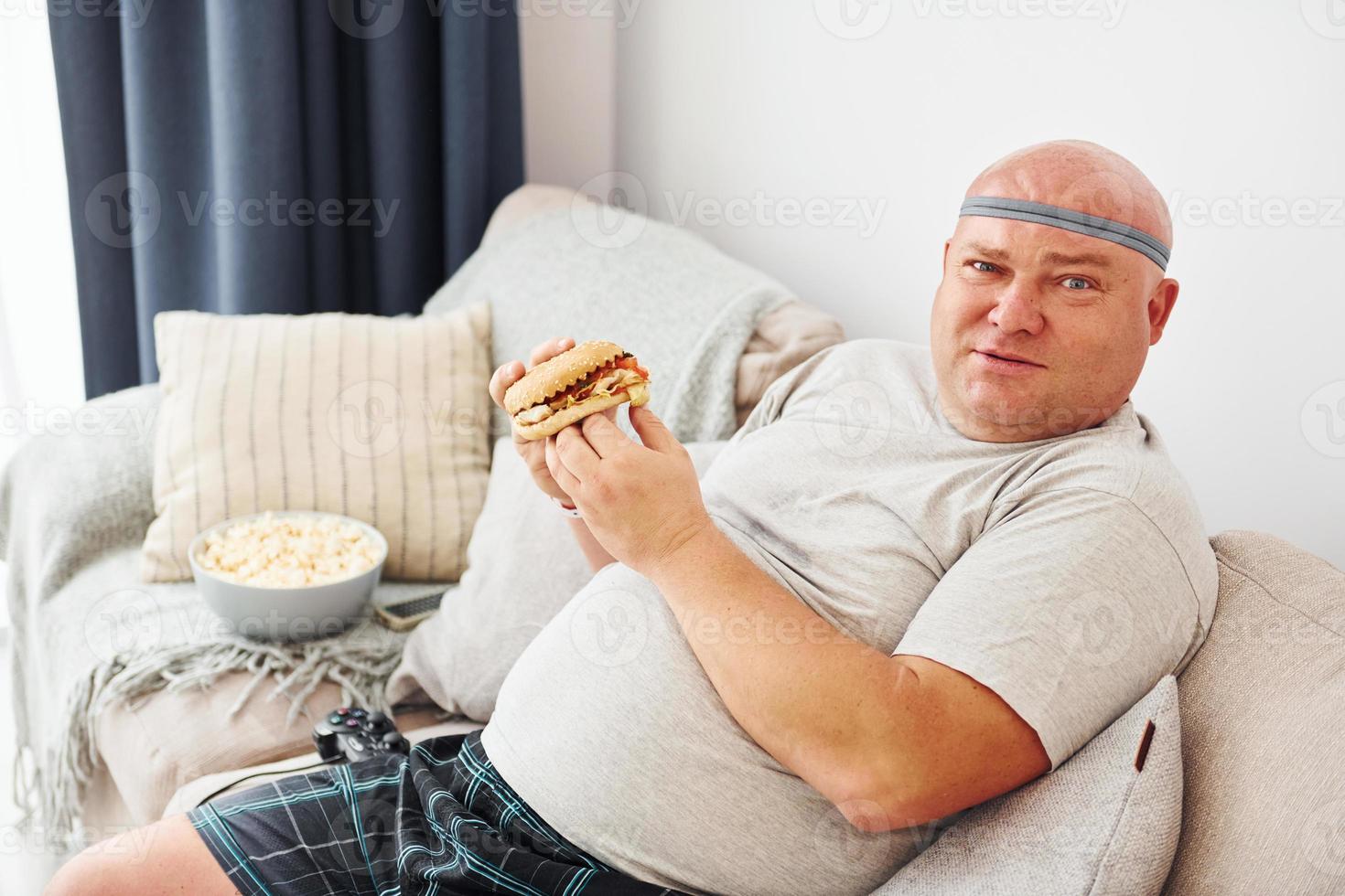 Sits on the sofa with popcorn and hamburger. Funny overweight man in casual clothes is indoors at home photo