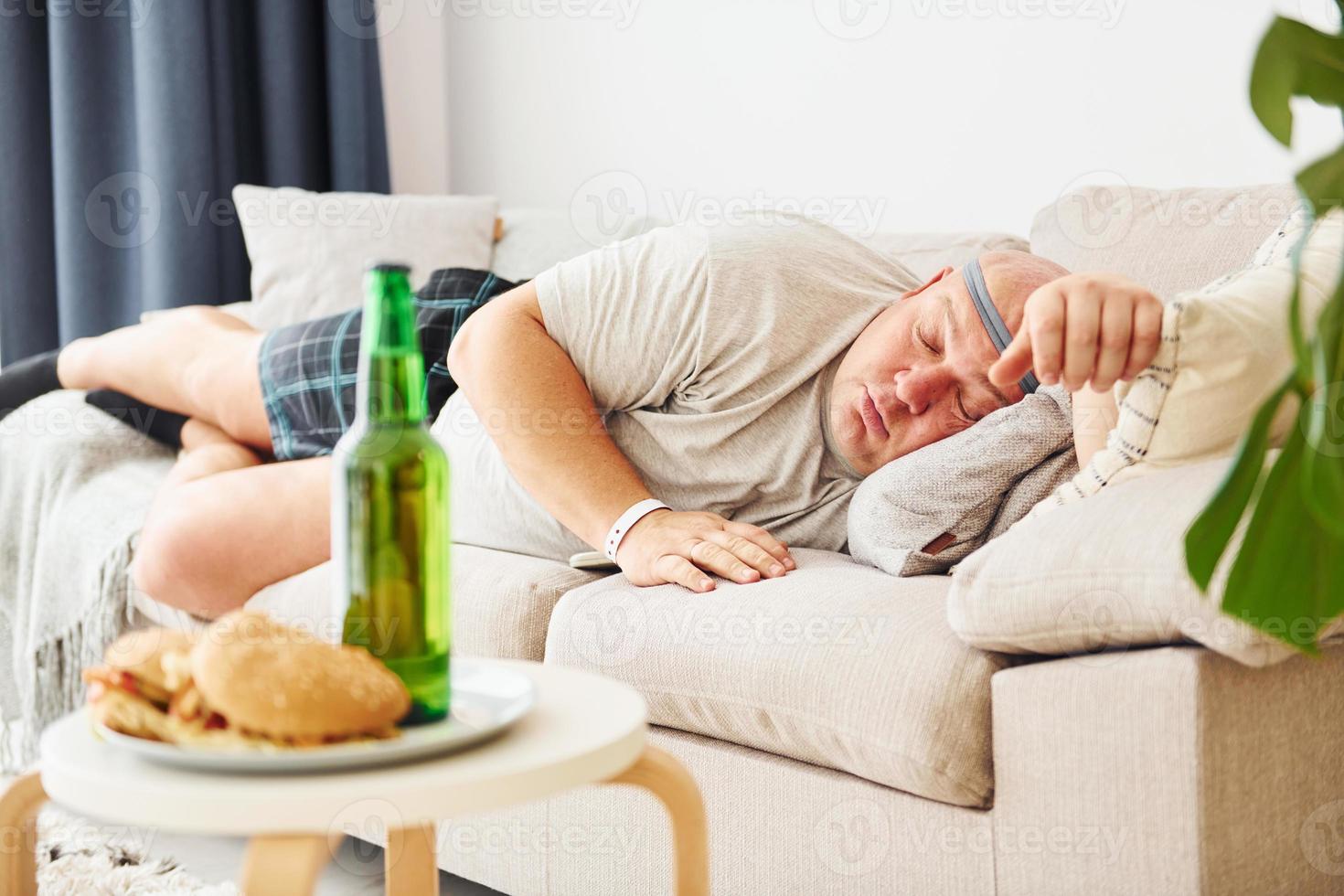 hamburguesas y cerveza. hombre acostado en el sofá. un hombre gracioso con sobrepeso vestido con ropa informal está en el interior de su casa foto