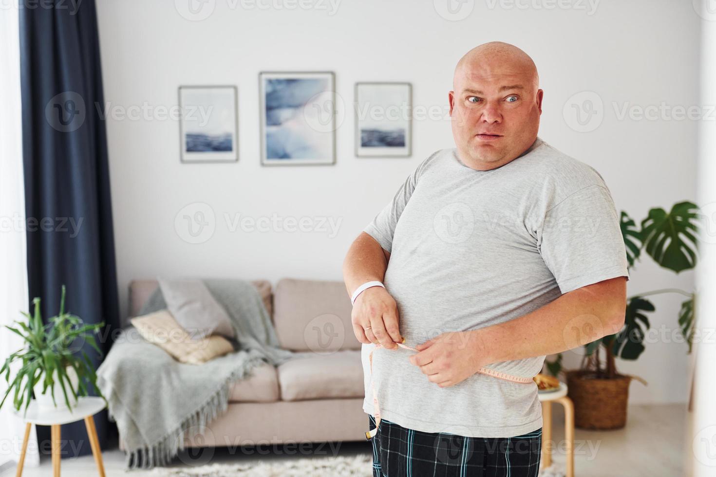 medir la cintura. un hombre gracioso con sobrepeso vestido con ropa informal está en el interior de su casa foto