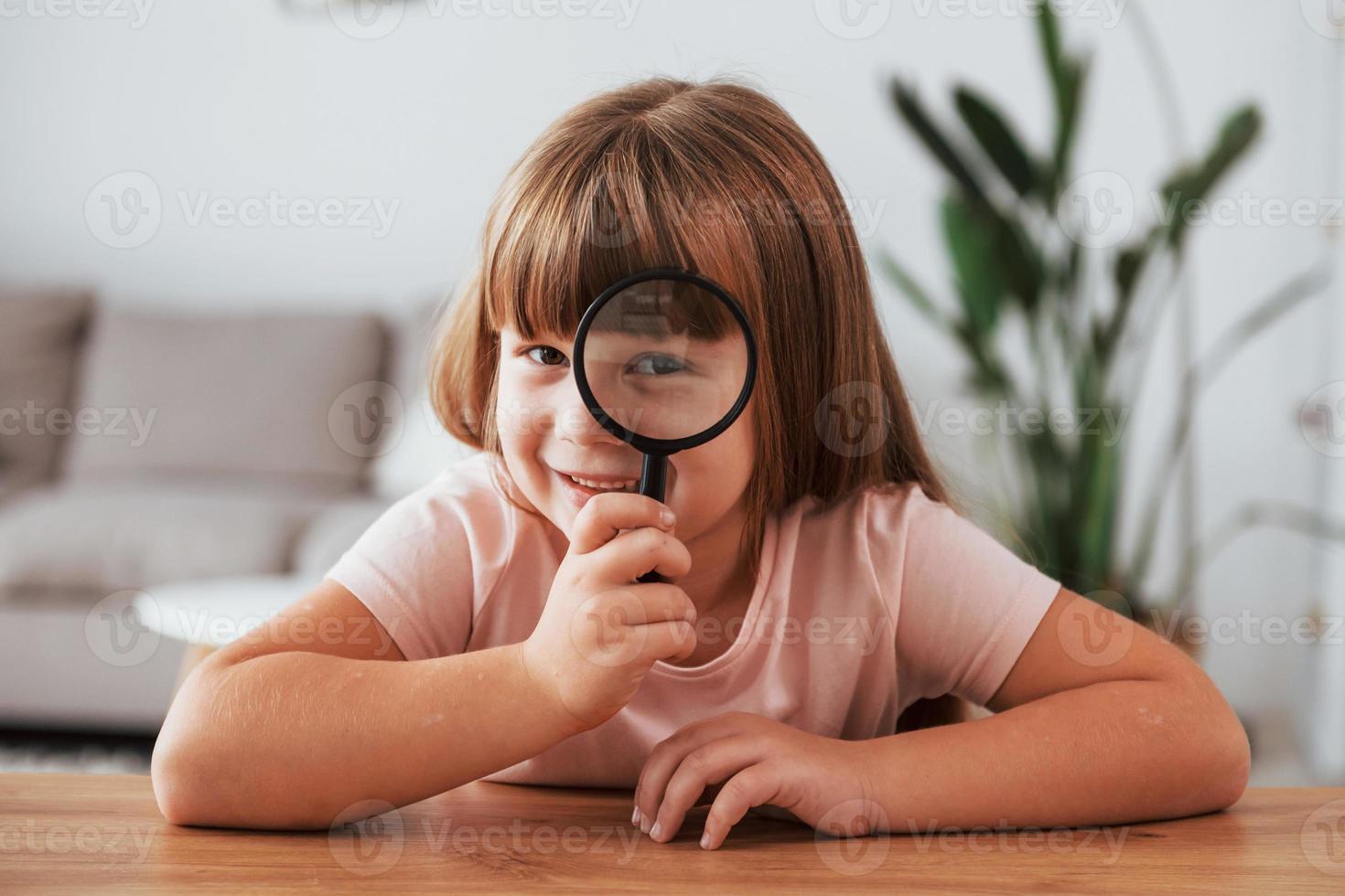 Having fun. Little girl is sitting by the table with magnifying glass photo